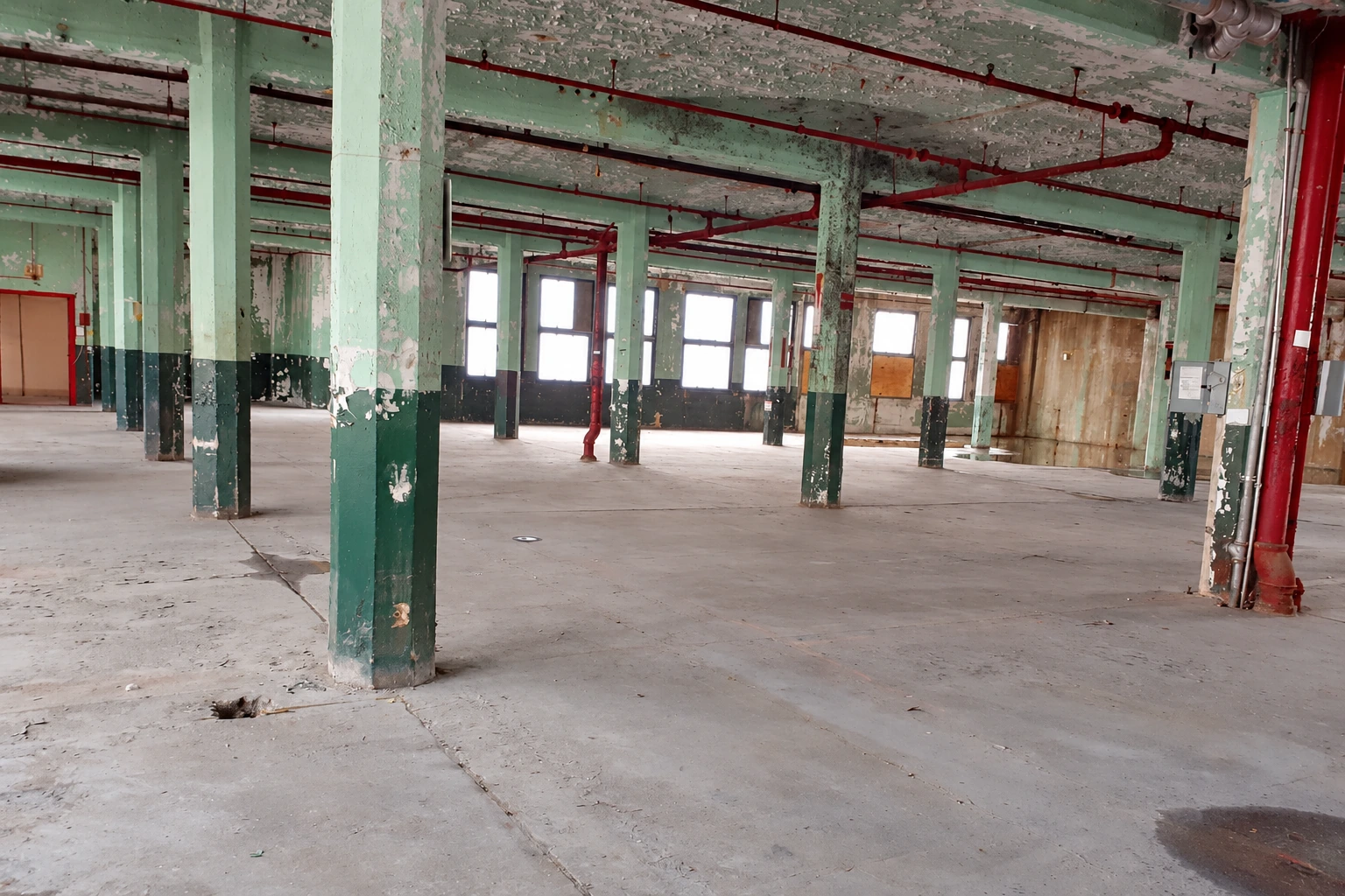 Existing industrial interior with green columns, exposed sprinkler piping and an unfinished concrete floor.